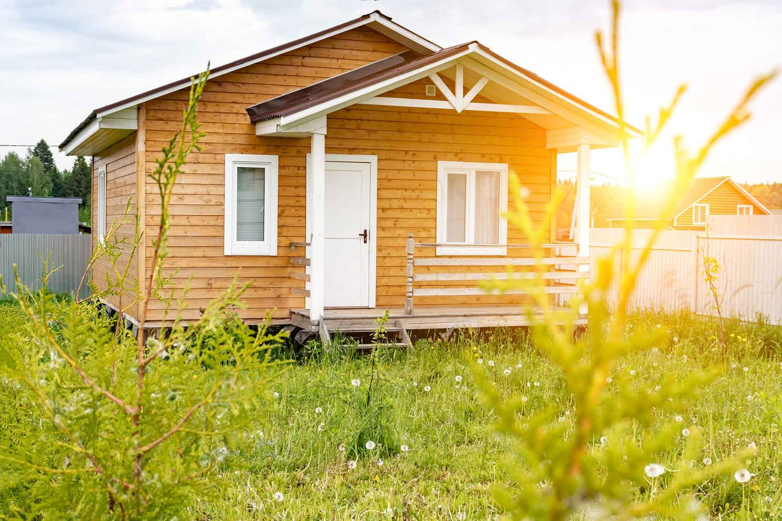 Petite maison en bois avec une façade blanche, baignée de lumière solaire, située dans un jardin verdoyant.