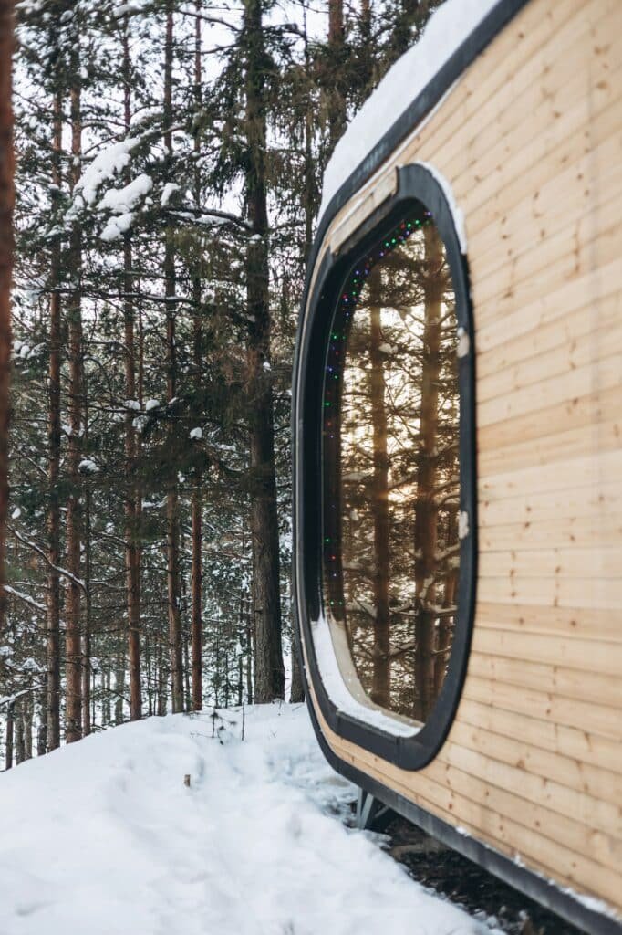 Vue latérale d'une tiny house en bois avec une grande fenêtre ovale, située dans une forêt enneigée.